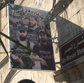 Kadunvarsikyltti jossa kolme miestä poseeraa kiväärien kanssa, ohessa arabiankielistä tekstiä.
