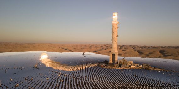 View of desert landscape with hundreds of solar panels surrounding a lighthouse-looking tower.