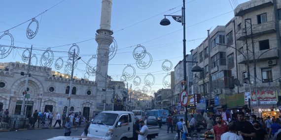 Kuvassa Ammanin kadulla kulkevaa ihmisjoukkoa ja liikennettä. Taustalla näkyy moskeijan minareetti sekä nousevan auringon valonsäteitä.