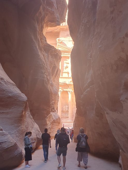 Kuvassa ihmisiä kävelemässä kapeaa kallliokanjonia pitkin kohti Petraa, jonka kallioon kaiverrettua fasadia näkyy kanjonin päässä.