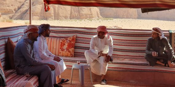 Kuvassa on neljä beduiinimiestä istuvat katetussa telttarakennuksessa aurinolta suojassa. Miehillä on valkoiset ja harmaat abayat päällään sekä päihinsä kiedotut ruudulliset huivit. He polttavat savukkeita ja juovat teetä.