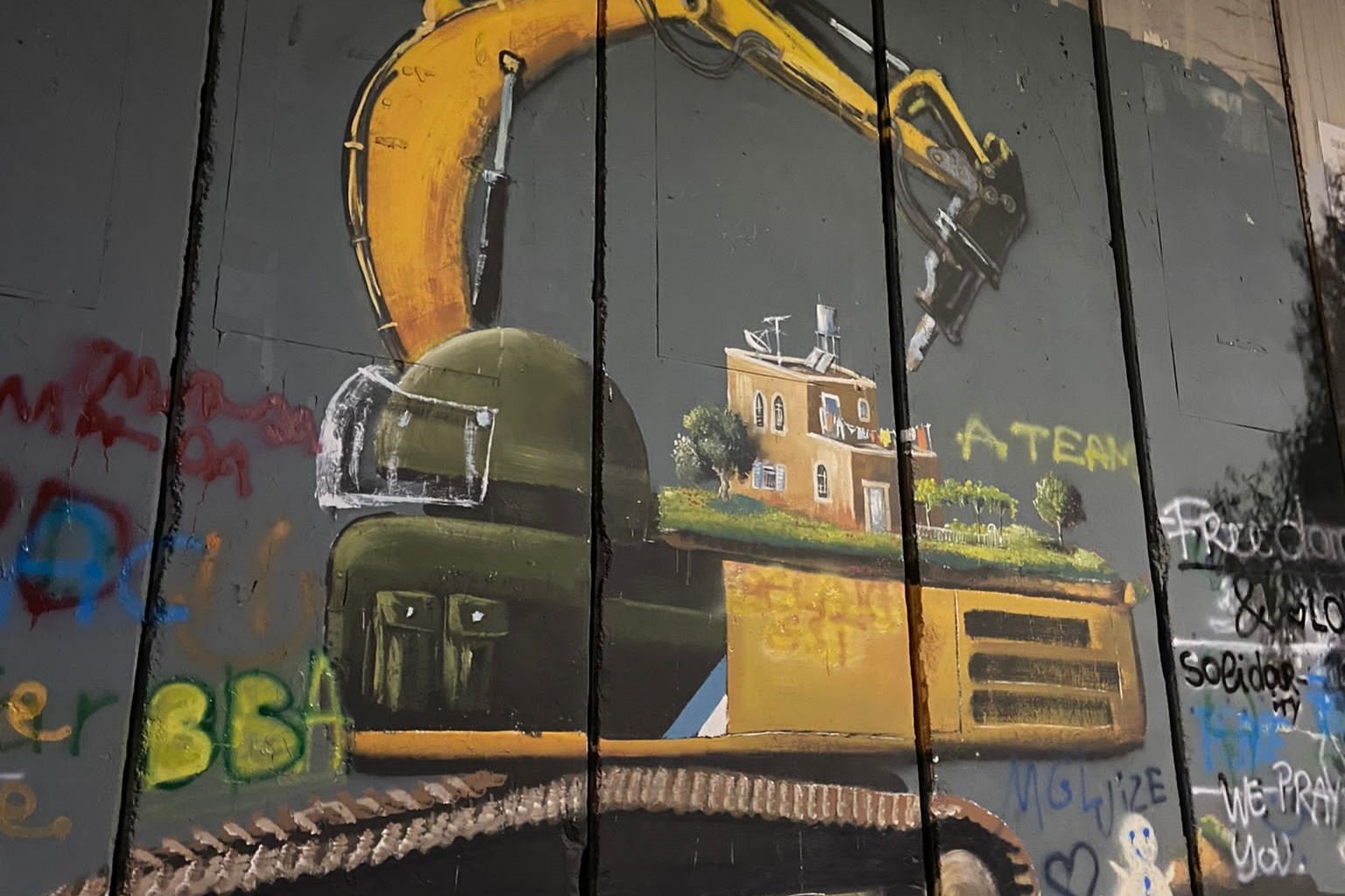 Graffiti keltaisesta paalutuskoneesta, jonka ohjaamo on kuvattu sotilaan kypäränä. Koneen selässä on ruksea, tasakattoinen asuintalo puutarhoineen, mitä koneen käsivarsi uhkaa.