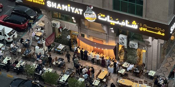 Kuvassa ylhäältäpäin ravintolan terassi, jolla useita, valkoisilla pöytäliinoilla peitettyjä pöytiä. Ravintolan ulkoseinustalla seisovapöytä, josta useat ihmiset ottavat ruokaa. Ihmisiä istuu myös pöytien äärellä. Ravintolalla on iso kelta-valkoinen valokyltti, jossa on sen nimi Shamiyat kirjoitettuna latinalaisin sekä arabialaisin aakkosin.