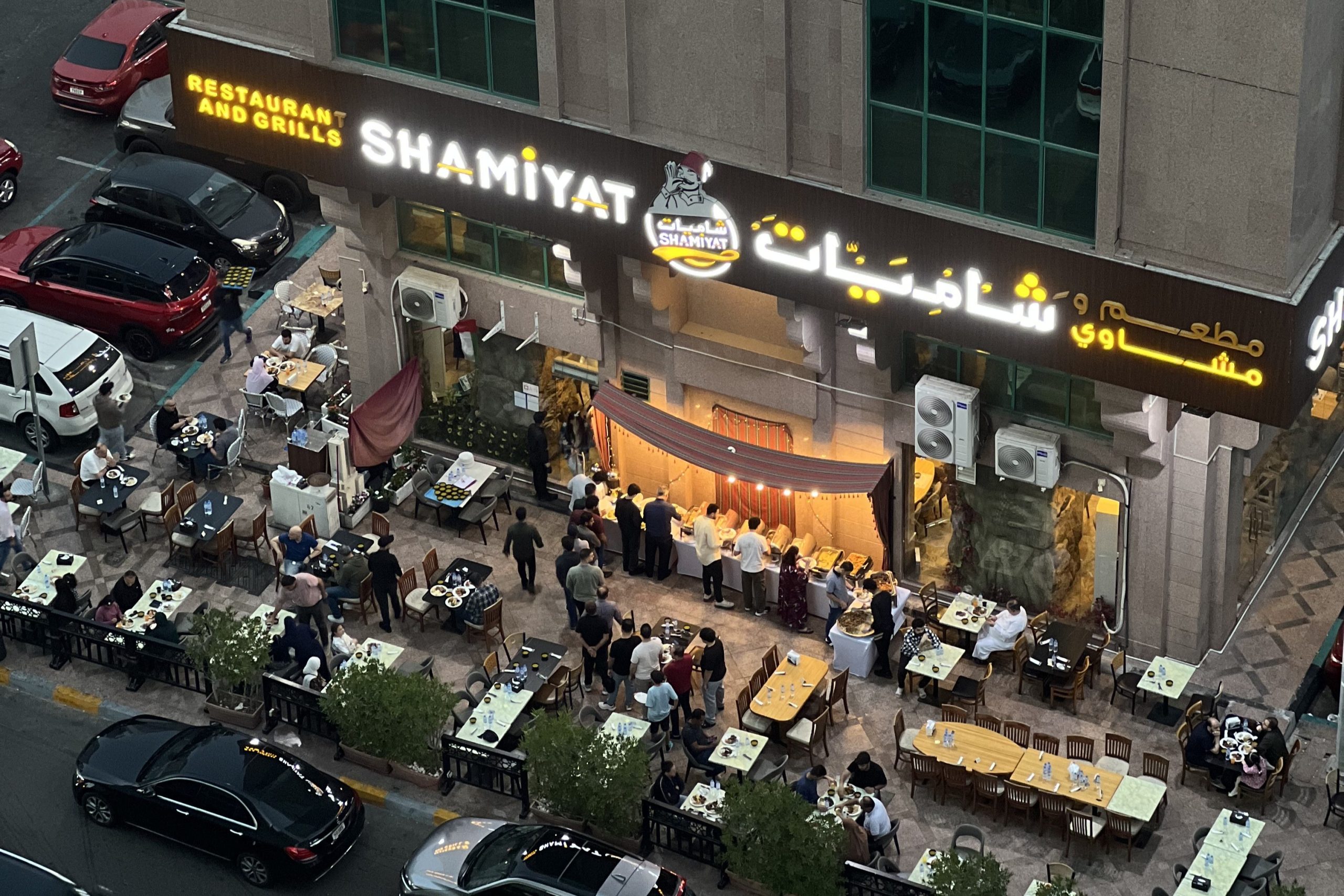 Kuvassa ylhäältäpäin ravintolan terassi, jolla useita, valkoisilla pöytäliinoilla peitettyjä pöytiä. Ravintolan ulkoseinustalla seisovapöytä, josta useat ihmiset ottavat ruokaa. Ihmisiä istuu myös pöytien äärellä. Ravintolalla on iso kelta-valkoinen valokyltti, jossa on sen nimi Shamiyat kirjoitettuna latinalaisin sekä arabialaisin aakkosin.