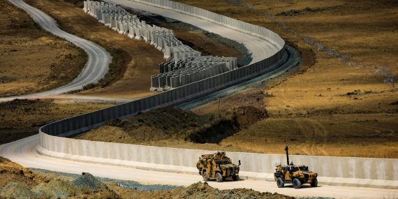Pitkä, mutkitteleva betoninen rajamuuri Turkin ja Iranin rajalla kulkee kuivassa, ruskeasävyisessä maastossa. Muurin vieressä kulkevalla huoltotielle ajaa kaksi panssaroitua sotilasajoneuvoa. Taustalla näkyy lisää betonisia esteitä ja kumpuilevaa, karua maisemaa.