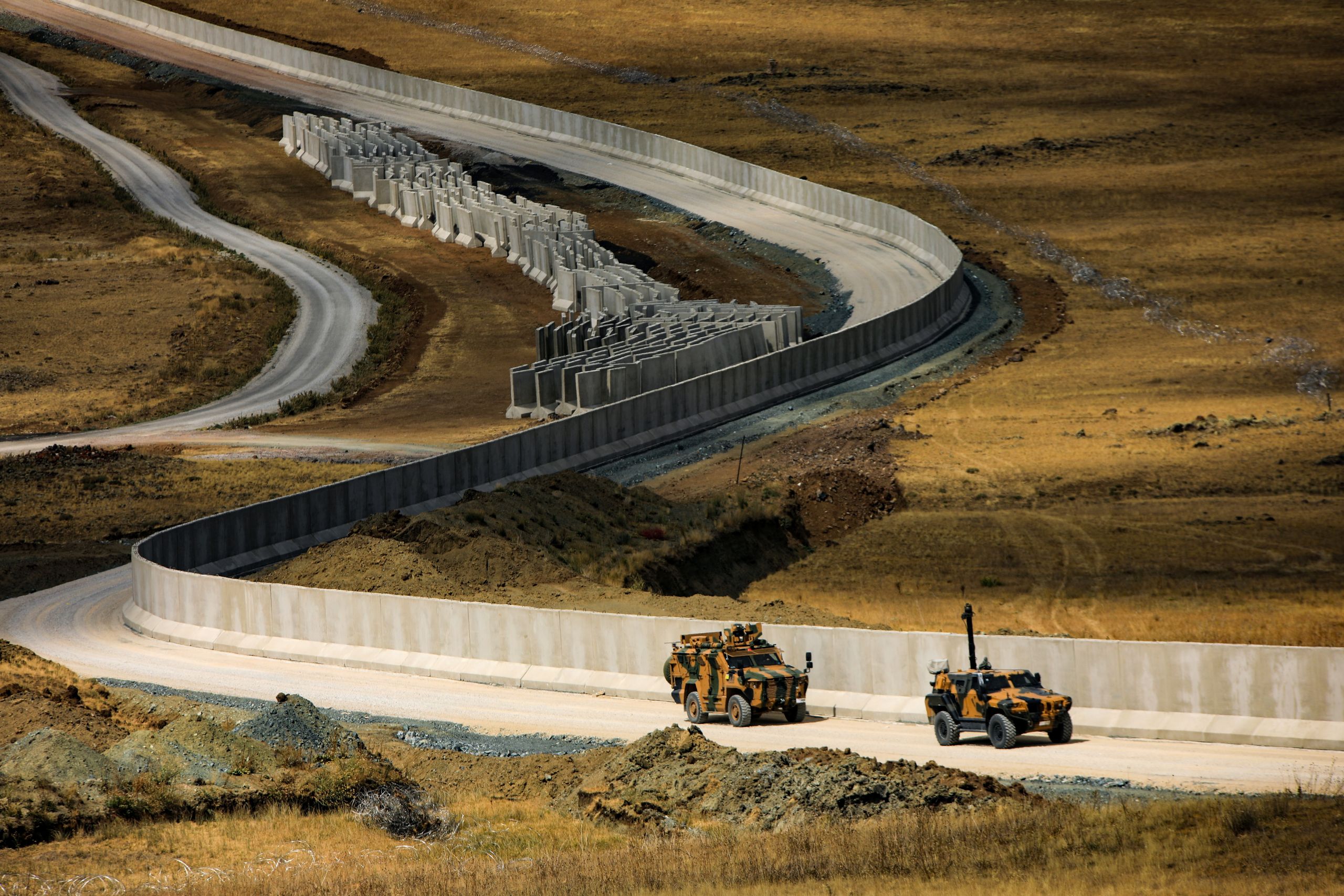 Pitkä, mutkitteleva betoninen rajamuuri Turkin ja Iranin rajalla kulkee kuivassa, ruskeasävyisessä maastossa. Muurin vieressä kulkevalla huoltotielle ajaa kaksi panssaroitua sotilasajoneuvoa. Taustalla näkyy lisää betonisia esteitä ja kumpuilevaa, karua maisemaa.
