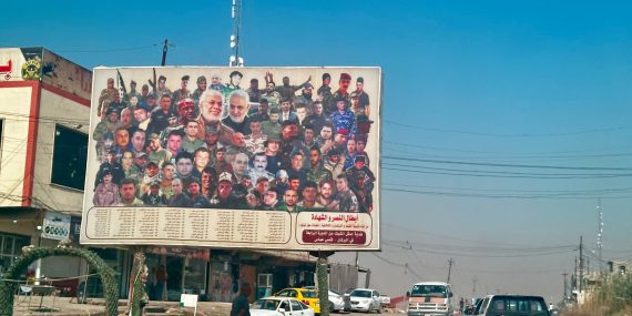 A large roadside billboard in Iraq showing a collage of dozens of men—many in military uniforms, with Arabic text listing names below, standing above a dusty road with cars, power lines, and low buildings.