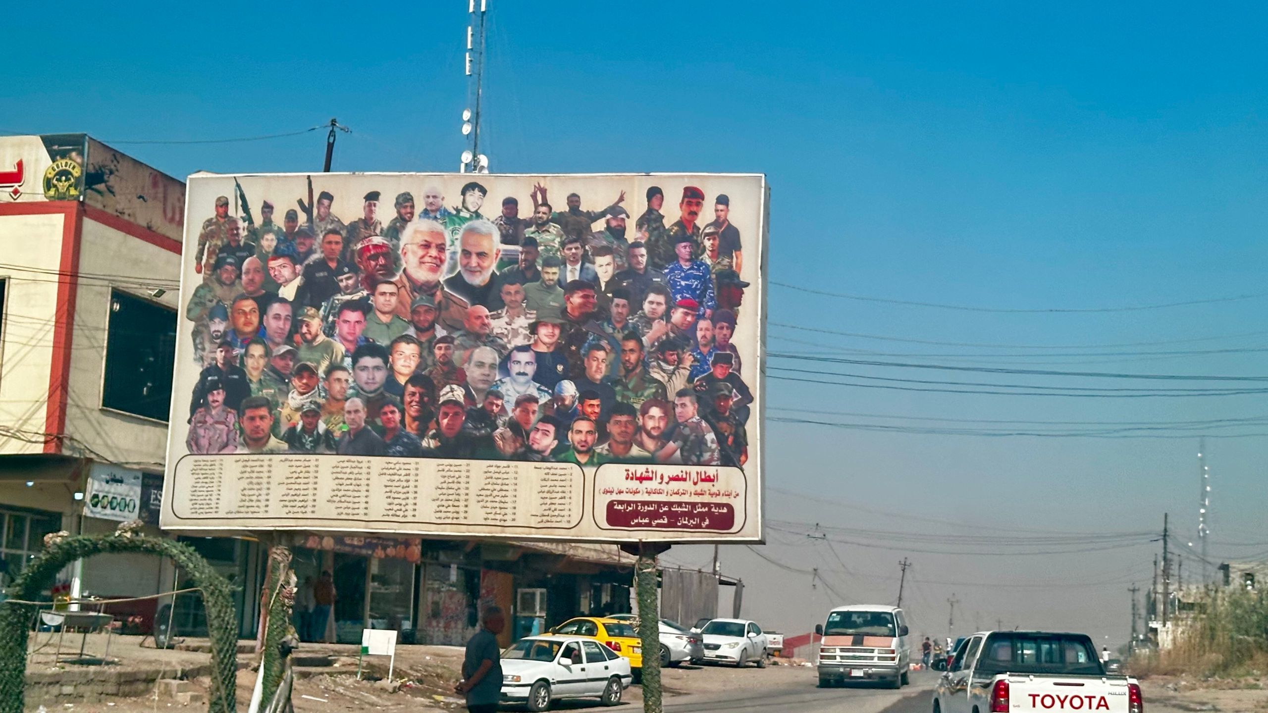 A large roadside billboard in Iraq showing a collage of dozens of men—many in military uniforms, with Arabic text listing names below, standing above a dusty road with cars, power lines, and low buildings.