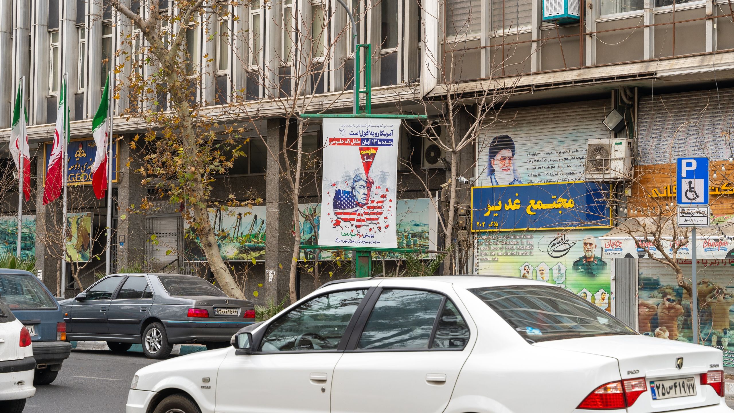 Kuvassa on katu Teheranissa Iranissa. Rakennuksen seinällä näkyy suuri poliittinen juliste, jossa Yhdysvaltain entinen presidentti Donald Trump on kuvattu Yhdysvaltain lipun väreissä, ja hänen yläpuolellaan on punainen nuolen tai tähtäimen kaltainen merkki. Julisteessa on persiankielistä tekstiä. Rakennuksen seinällä on myös suuri kuva Iranin uskonnollisesta johtajasta Ali Khameneista sekä muita poliittisia ja yhteiskunnallisia julisteita. Rakennuksen edessä liehuu useita Iranin lippuja. Kadulla kulkee autoja ja puita kasvaa jalkakäytävän varrella.