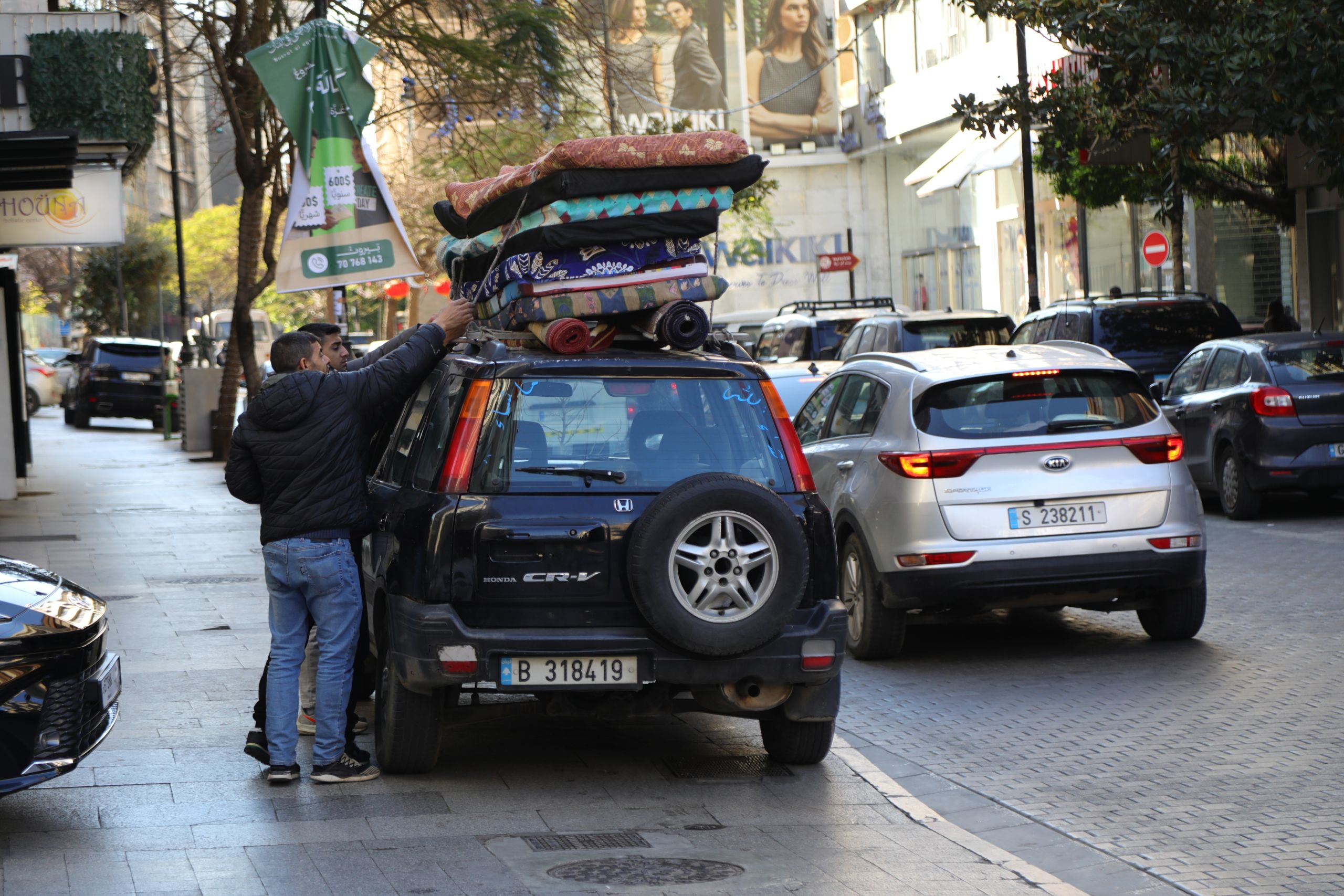 Kaksi miestä kiinnittää kuormaa auton katolle vilkkaalla kaupunkikadulla. Mustan katumaasturin katolla on korkea pino patjoja ja kankaita, joita miehet sitovat paikoilleen, samalla kun viereisellä kaistalla kulkee autoja. Taustalla näkyy puita, liikkeitä ja mainoskylttejä.