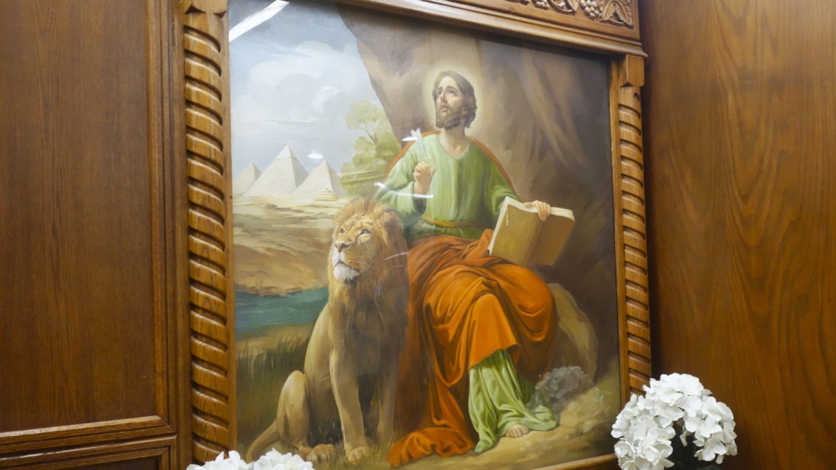 Kehystetty maalaus Pyhästä Markuksesta vihreässä ja punaisessa asussa istumassa leijonan vieressä. Hän katsoo ylöspäin ja pitää Raamattua kädessään, taustalla näkyy pyramideja ja maisemaa.