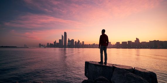 Mies seisoo kallioilla veden äärellä katsomassa Abu Dhabin siluettia auringonnousussa; taivas hehkuu pinkin ja oranssin sävyissä ja pilvenpiirtäjät piirtyvät horisonttiin.
