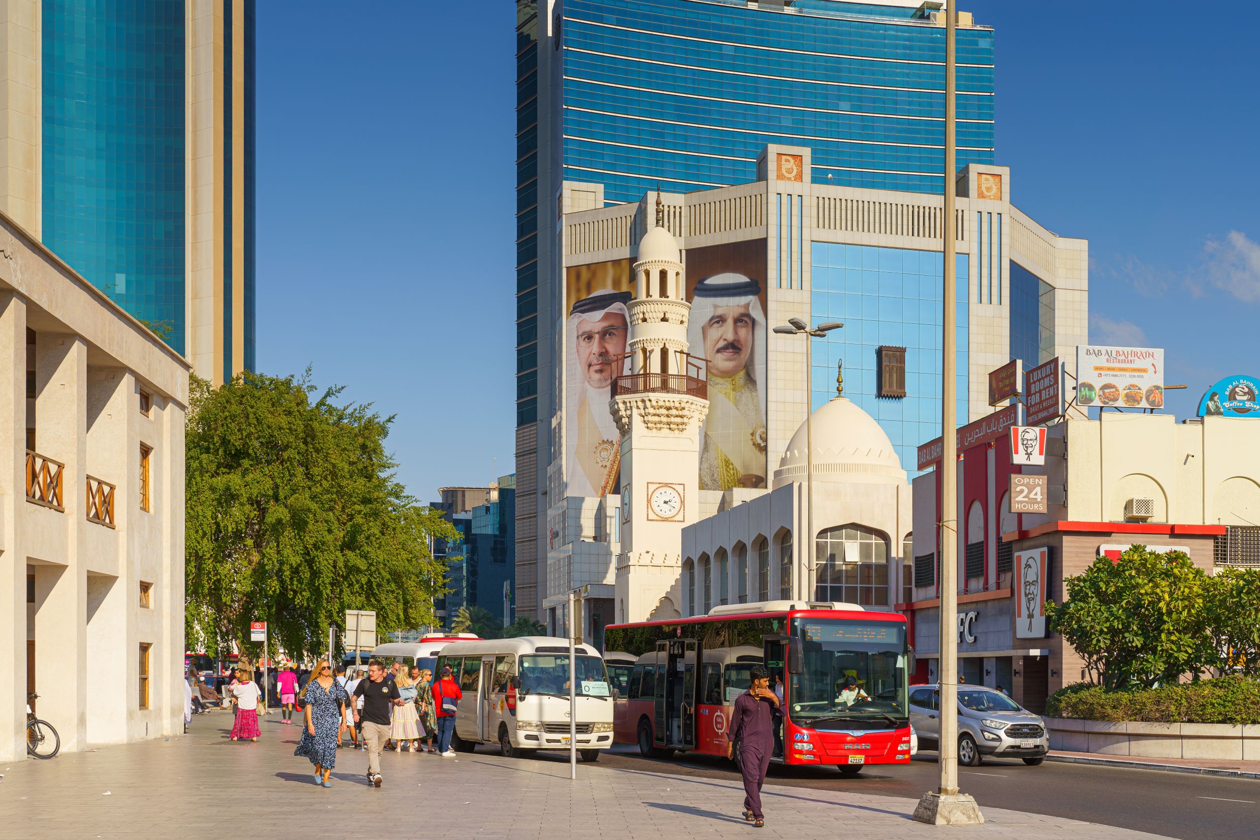 Aurinkoinen katunäkymä Bahrainista: jalankulkijoita kävelee leveällä jalkakäytävällä ja kadulla kulkee busseja. Taustalla kohoaa moderni lasinen toimistorakennus, jonka julkisivussa on suuret muotokuvat emiiristä ja kruununprinssistä. Etualalla näkyy pieni moskeija kupoleineen ja minareetti, ja oikealla puolella on ravintoloita sekä liiketiloja.