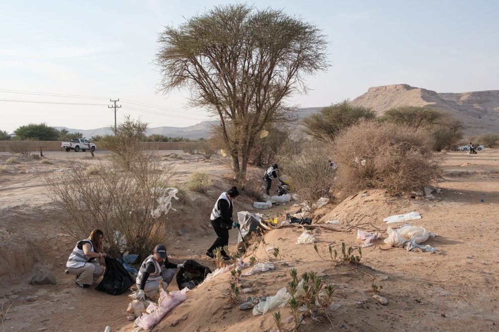 Viisi vapaaehtoista kerää roskia kuivassa autiomaamaisemassa Saudi-Arabiassa. He käyttävät hanskoja ja keräävät muovijätettä maasta pusseihin pensaiden ja puun ympärillä. Taustalla näkyy tie, auto sekä matalia vuoria.