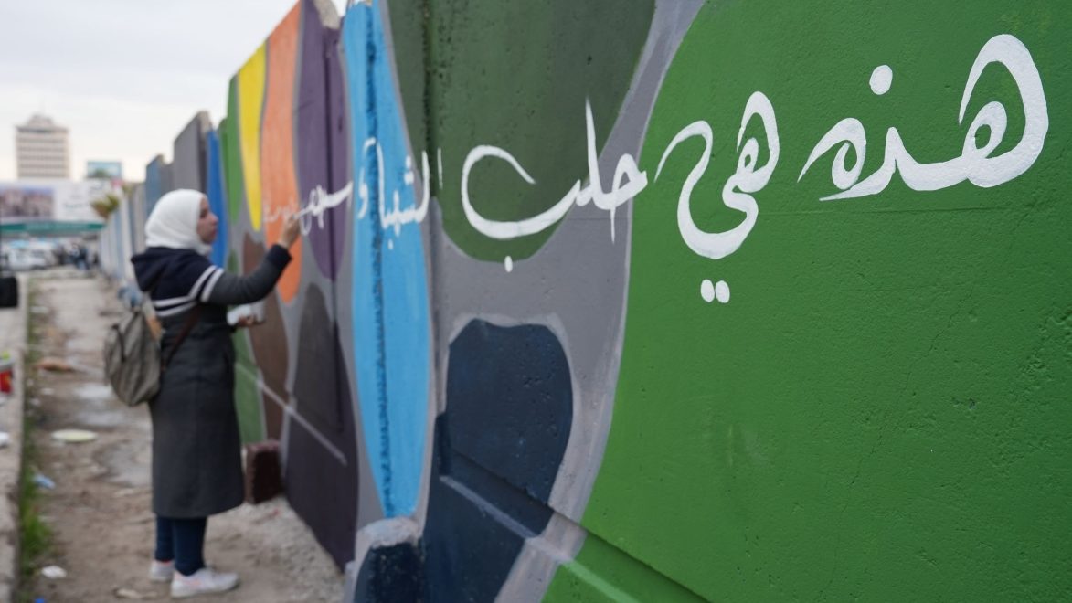 Nainen maalaa värikästä graffitia ulkona. Etualalla vihreään seinään on maalattu suurikokoista valkoista arabialaista kalligrafiaa, ja taustalla näkyy muita värikkäitä seinäpintoja sekä kaupunkimaisemaa.