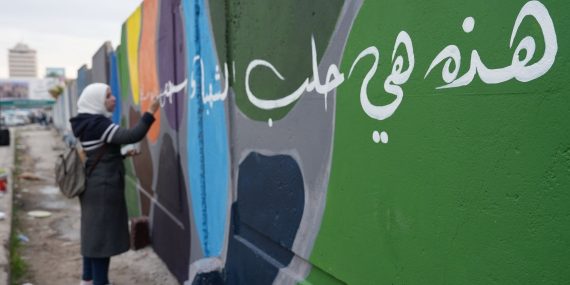 Nainen maalaa värikästä graffitia ulkona. Etualalla vihreään seinään on maalattu suurikokoista valkoista arabialaista kalligrafiaa, ja taustalla näkyy muita värikkäitä seinäpintoja sekä kaupunkimaisemaa.