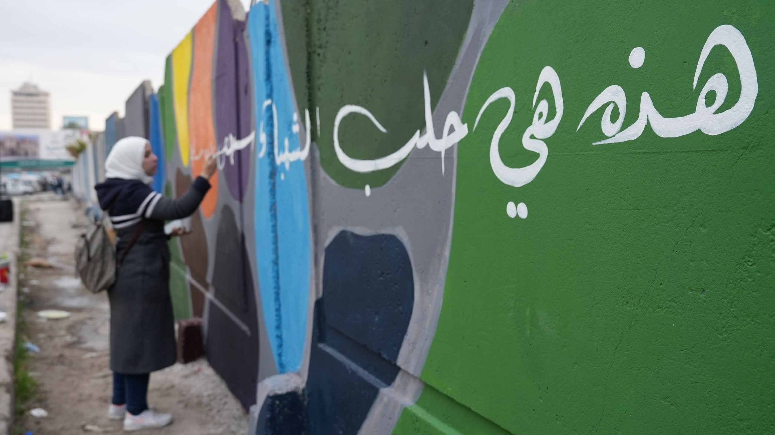 Nainen maalaa värikästä graffitia ulkona. Etualalla vihreään seinään on maalattu suurikokoista valkoista arabialaista kalligrafiaa, ja taustalla näkyy muita värikkäitä seinäpintoja sekä kaupunkimaisemaa.