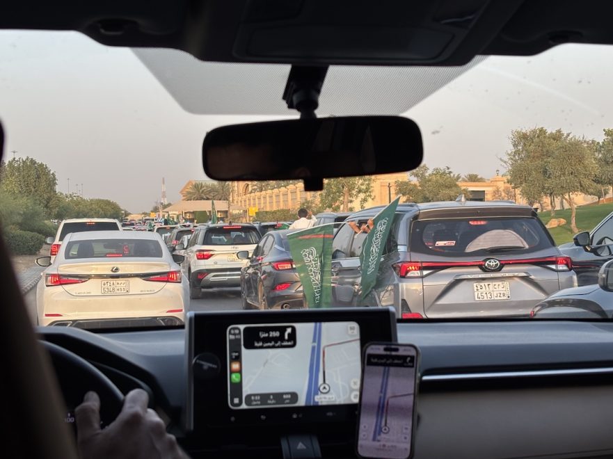 Auton etuikkunasta näkyvä ruuhkainen tie ja autojono. Autojen ikkunoista heiluu Saudi-Arabian lippuja. 