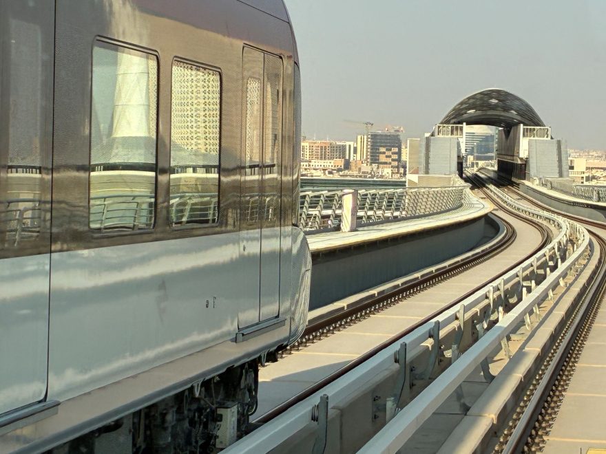 Riadin metrorata päiväsaikaan. Vasemmalla näkyy metron vaunu, joka kaartuu viereisen raiteen ohi. Edessä raide kaartuu moderniin, kupolimaiseen metroasemarakennukseen, ja taustalla näkyy Riadin kaupunkimaisemaa.