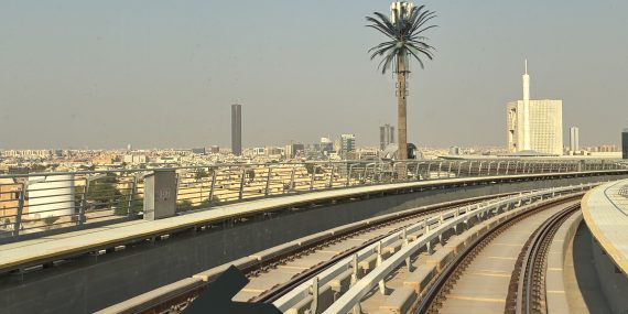 Näkymä Riadin kaupunkiin metroradalta. Etualalla kaartuvat metroradat ja aseman rakenteita, taustalla laaja kaupunkimaisema. Keskellä rataa reunustaa korkea palmu, ja oikealla näkyy suuri moderni rakennus.